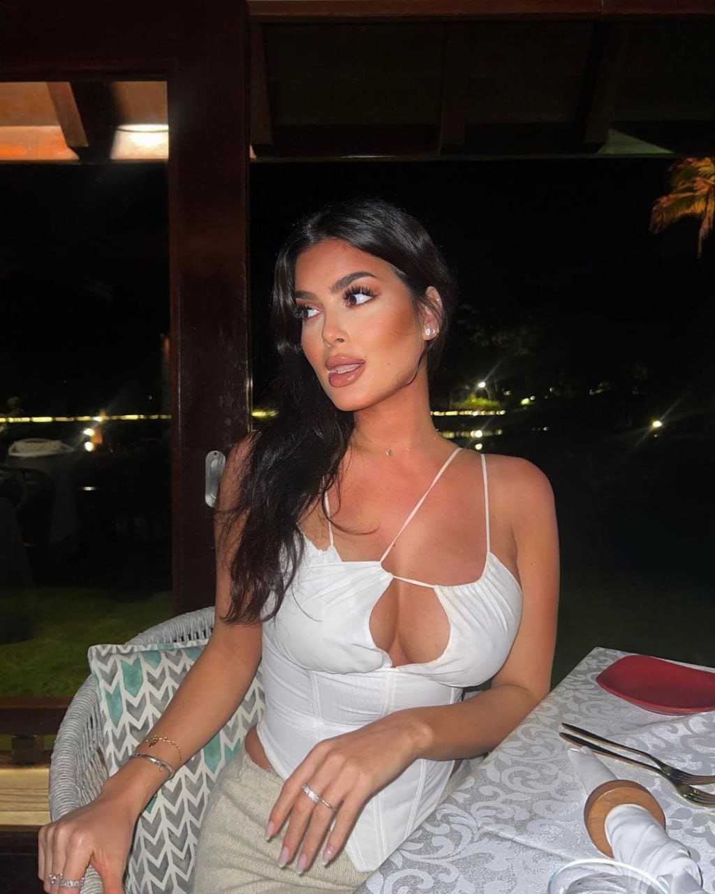 Mirabella sits at a table outdoors at night, wearing a white corset top with a keyhole cutout and a light skirt against a dark background.
