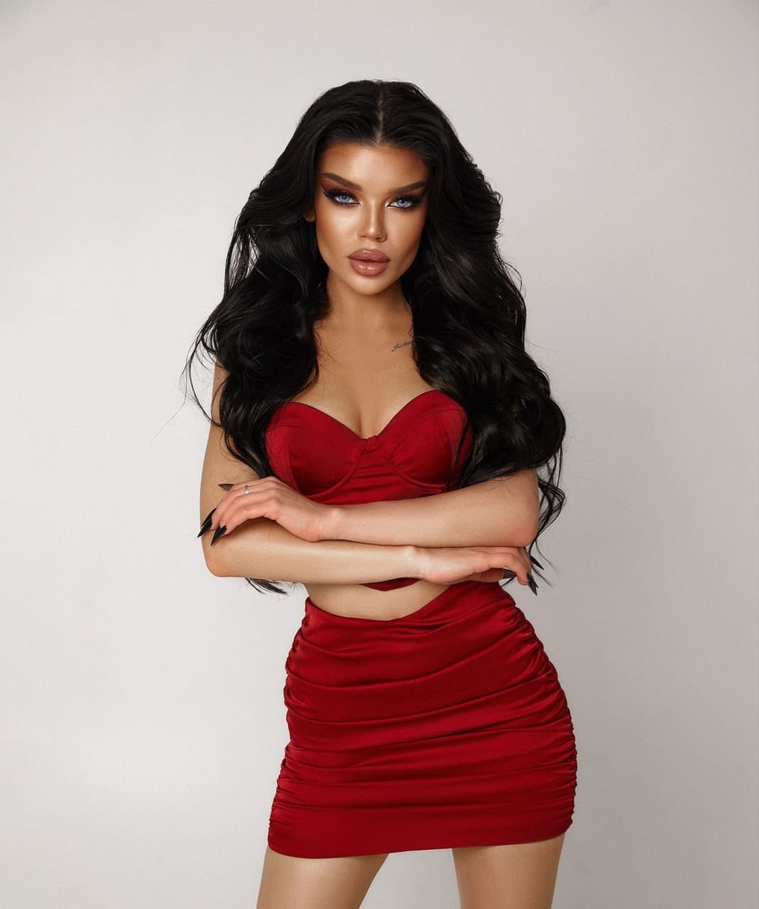 Jasmine stands against a light background in a red satin strapless crop top and ruched mini skirt, arms crossed over her midriff.