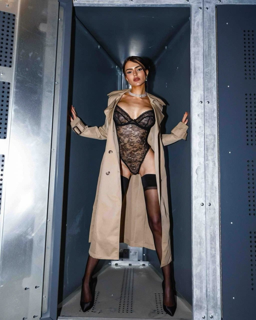 Woman standing in a metallic ventilation shaft wearing an open beige trench coat, black lace bodysuit, and stockings.
