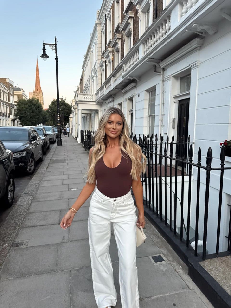 Angela walks down a city sidewalk in a dark maroon sleeveless bodysuit with a deep scoop neckline and white high-waisted trousers.