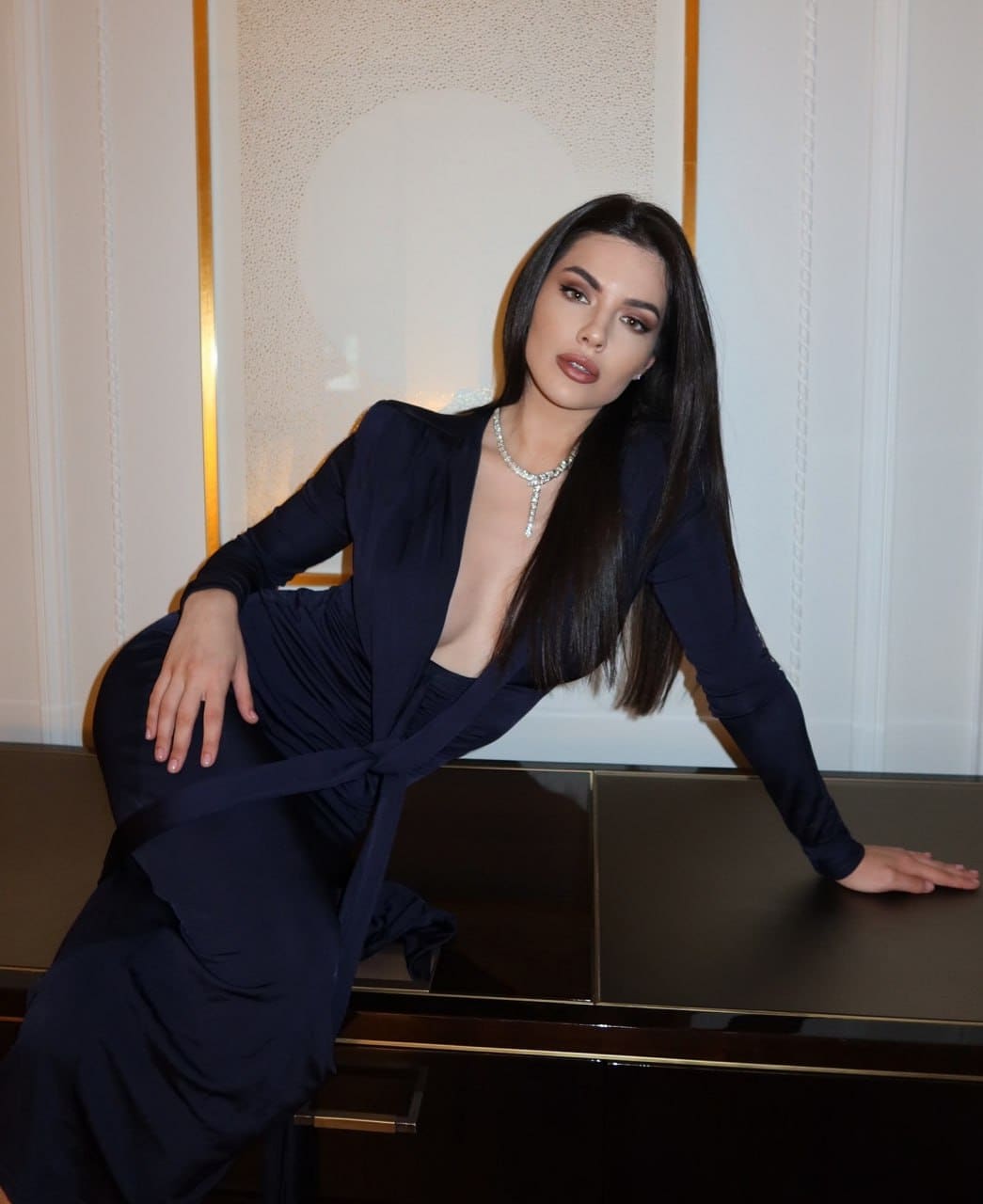 Lydia leans back against a dark surface in a floor-length navy gown with long sleeves, a plunging neckline, and a diamond necklace.