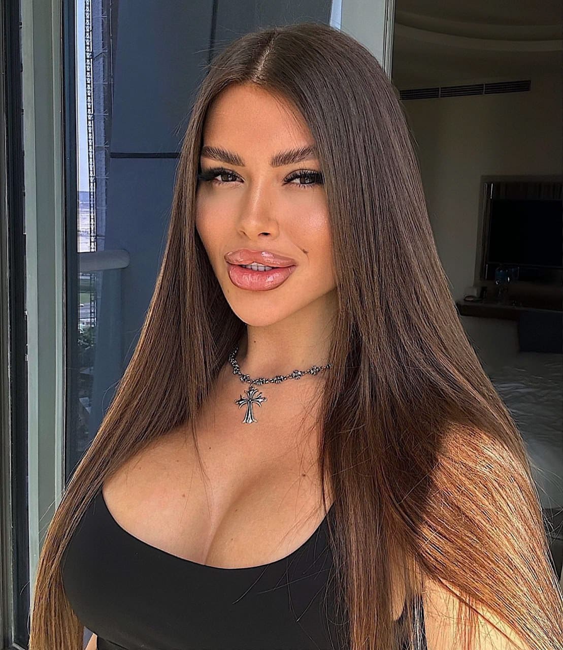 A woman with long dark hair poses indoors in a black top with a deep scoop neckline and a silver cross necklace.