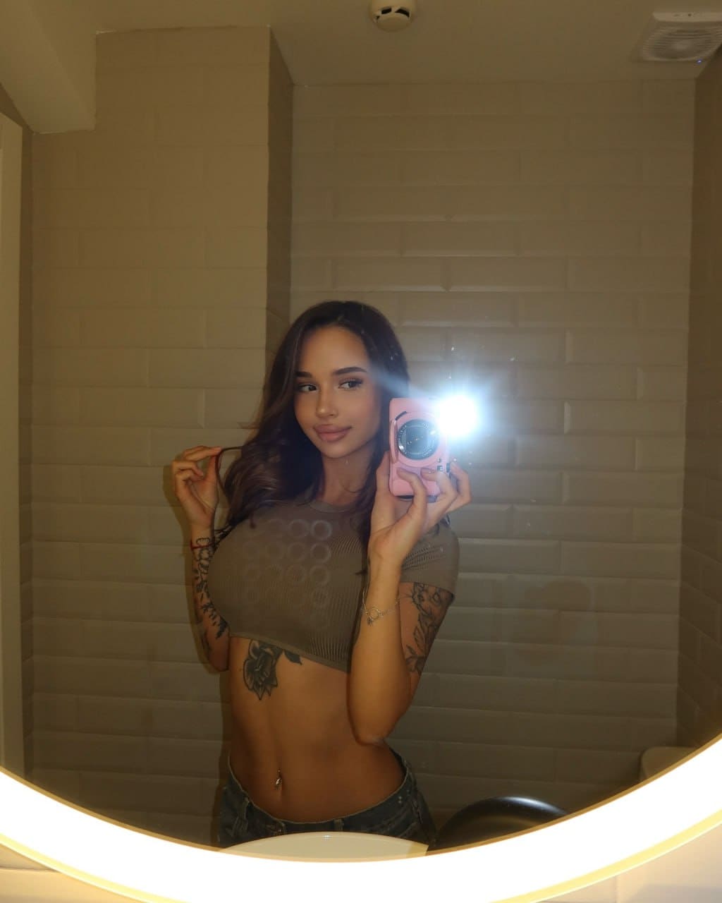 Mirror selfie with a pink camera in an olive green crop top and denim shorts, revealing a navel piercing against white brick.