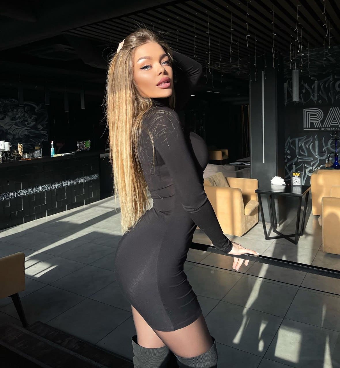 Woman stands in profile in a lounge wearing a tight black mini dress and thigh-high boots, hand resting on her head.