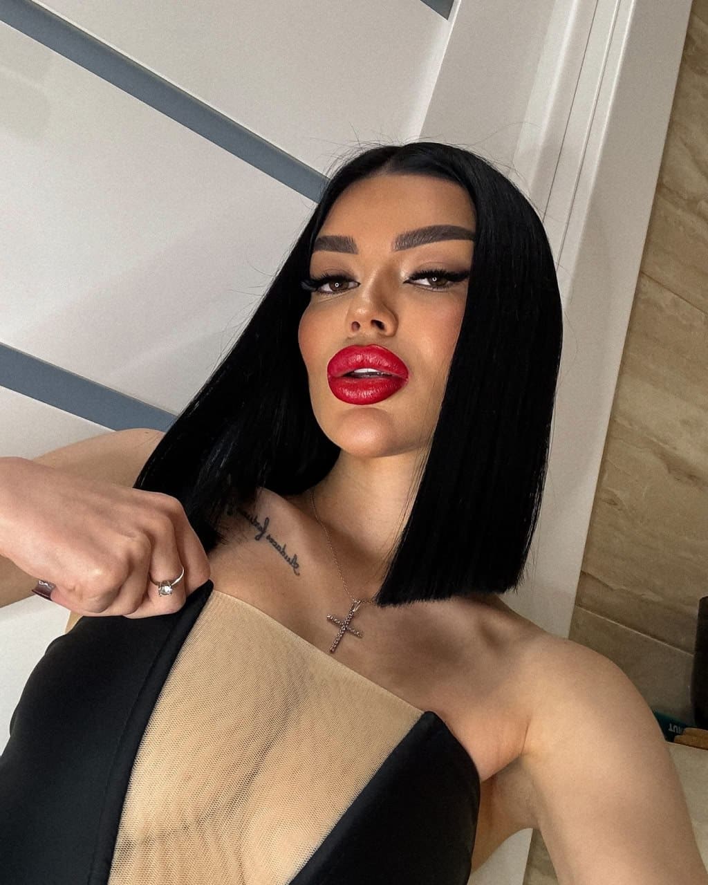 A woman with a black bob and red lipstick wears a sheer mesh top and cross necklace, posing near a white door indoors.