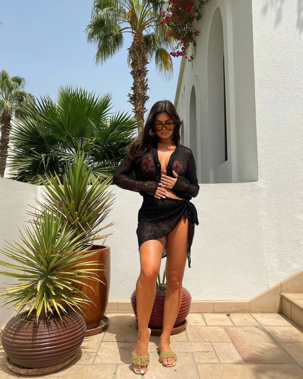 A woman in sunglasses stands on a tiled patio, wearing a sheer black open-knit cardigan and wrap skirt over a bikini, with embellished sandals.