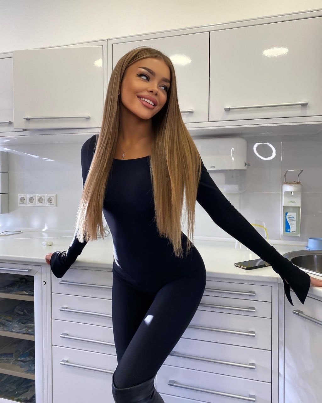 Halia leans back against a white kitchen counter in a black long-sleeved bodysuit, leggings, and tall black boots.