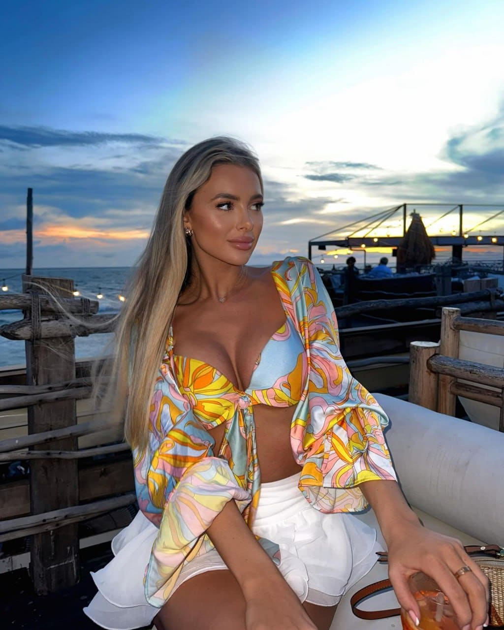 Woman sits outdoors at dusk by a wooden railing, holding a drink, wearing a colorful patterned top with bell sleeves and a white skirt.