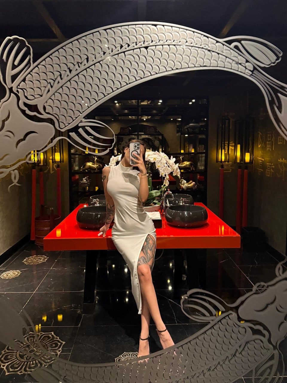 Leaning on a red table in a mirror selfie, wearing a white high-neck dress with a thigh slit revealing a leg tattoo.