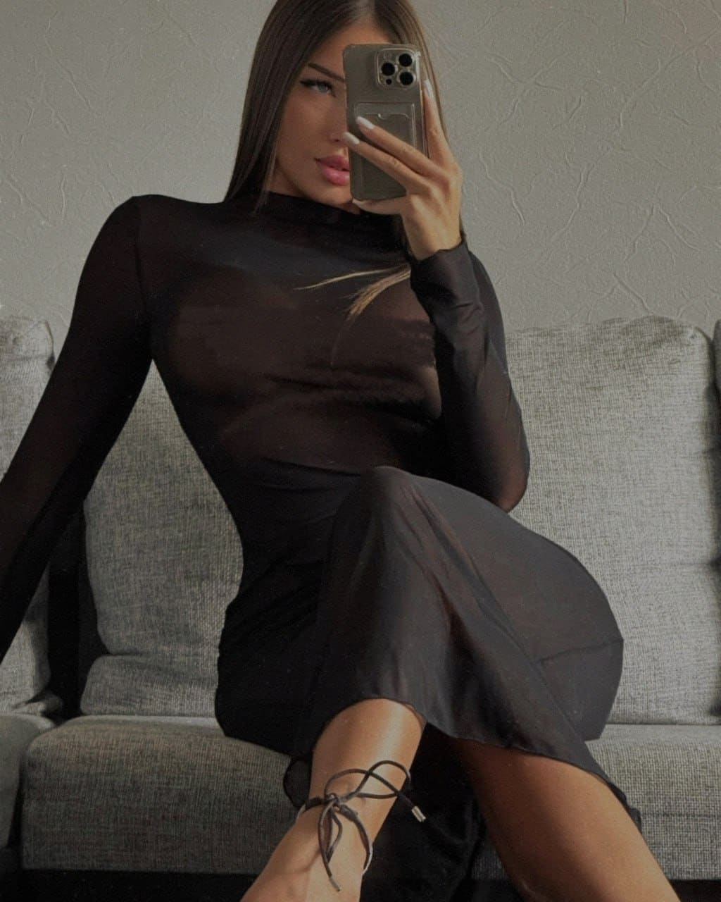 A woman sits on a gray sofa in a sheer black midi dress and strappy heels, taking a mirror selfie that reveals undergarment silhouettes.