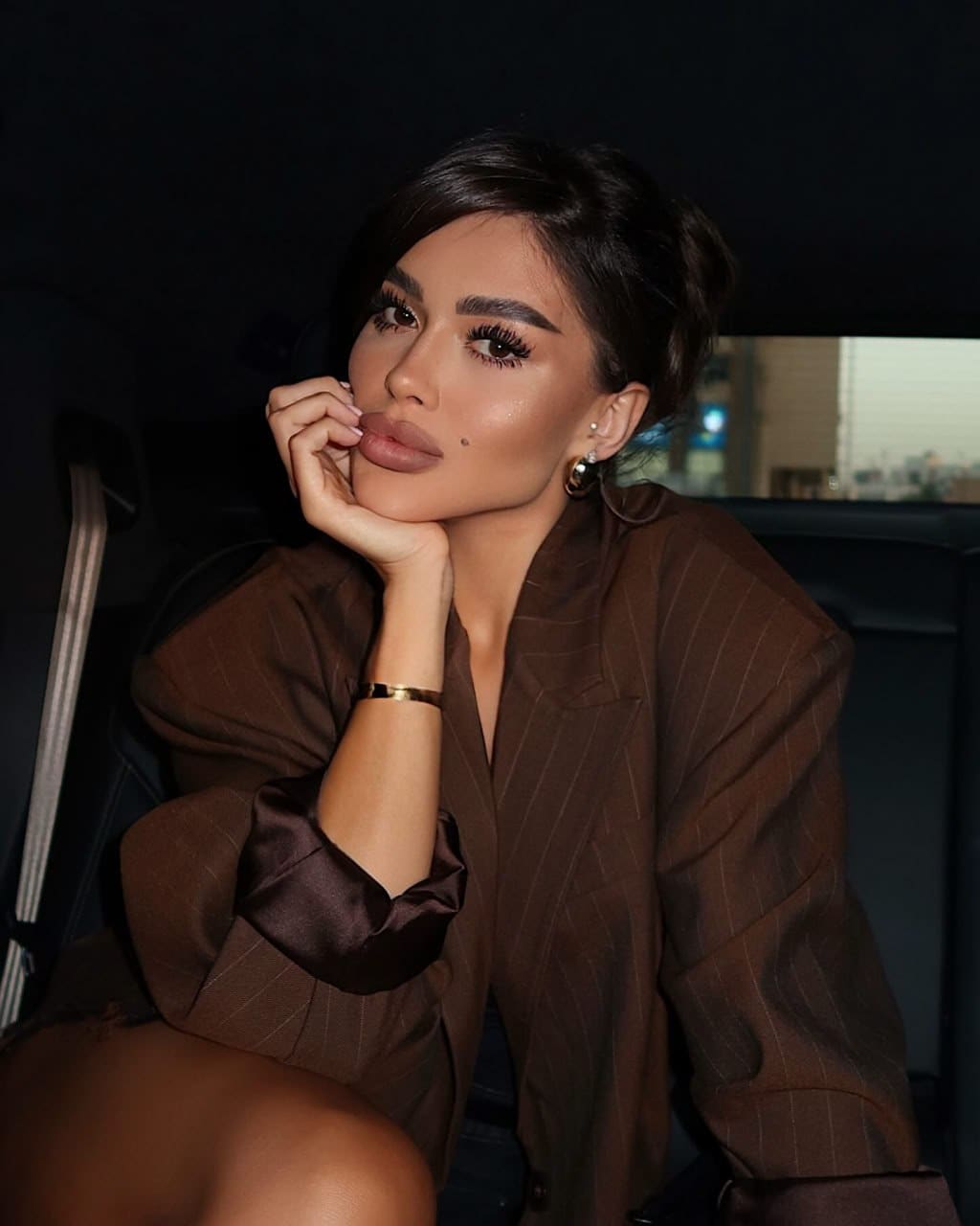 Kiki sits in a car resting her chin on her hand, wearing a brown pinstripe blazer with satin cuffs and hoop earrings.
